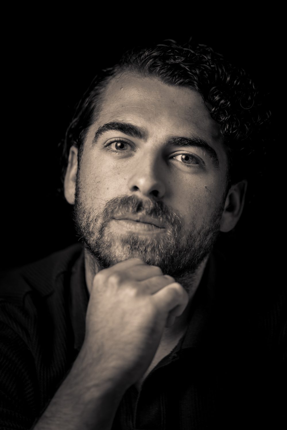 Zwart-witportret van een man met krullend haar die zijn kin op zijn vuist laat rusten, tegen een zwarte achtergrond
